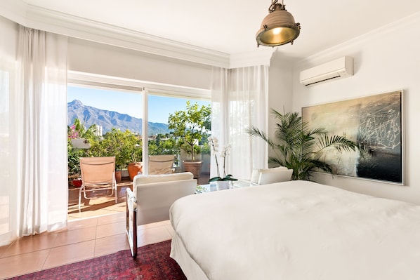 A hotel room with a large bed, a desk, and a chair. There is a view of mountains and a balcony with plants.