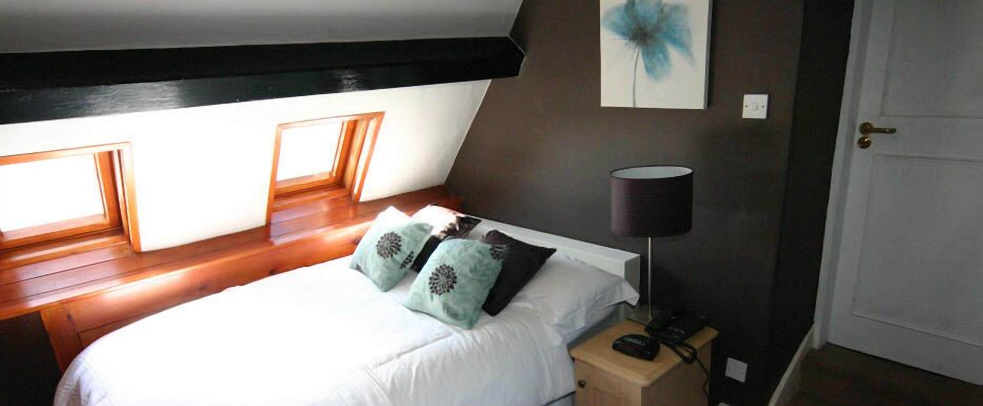 A bedroom with a sloped ceiling, a bed with white bedding and patterned pillows, a bedside lamp, and a door.