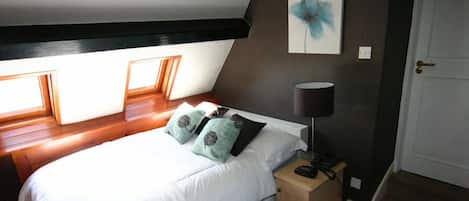 A bedroom with a sloped ceiling, a bed with white bedding and patterned pillows, a bedside lamp, and a door.