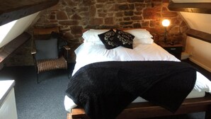 A cozy room with a stone wall, a bed with white bedding and black pillows, a small chair, and a table with a lamp.