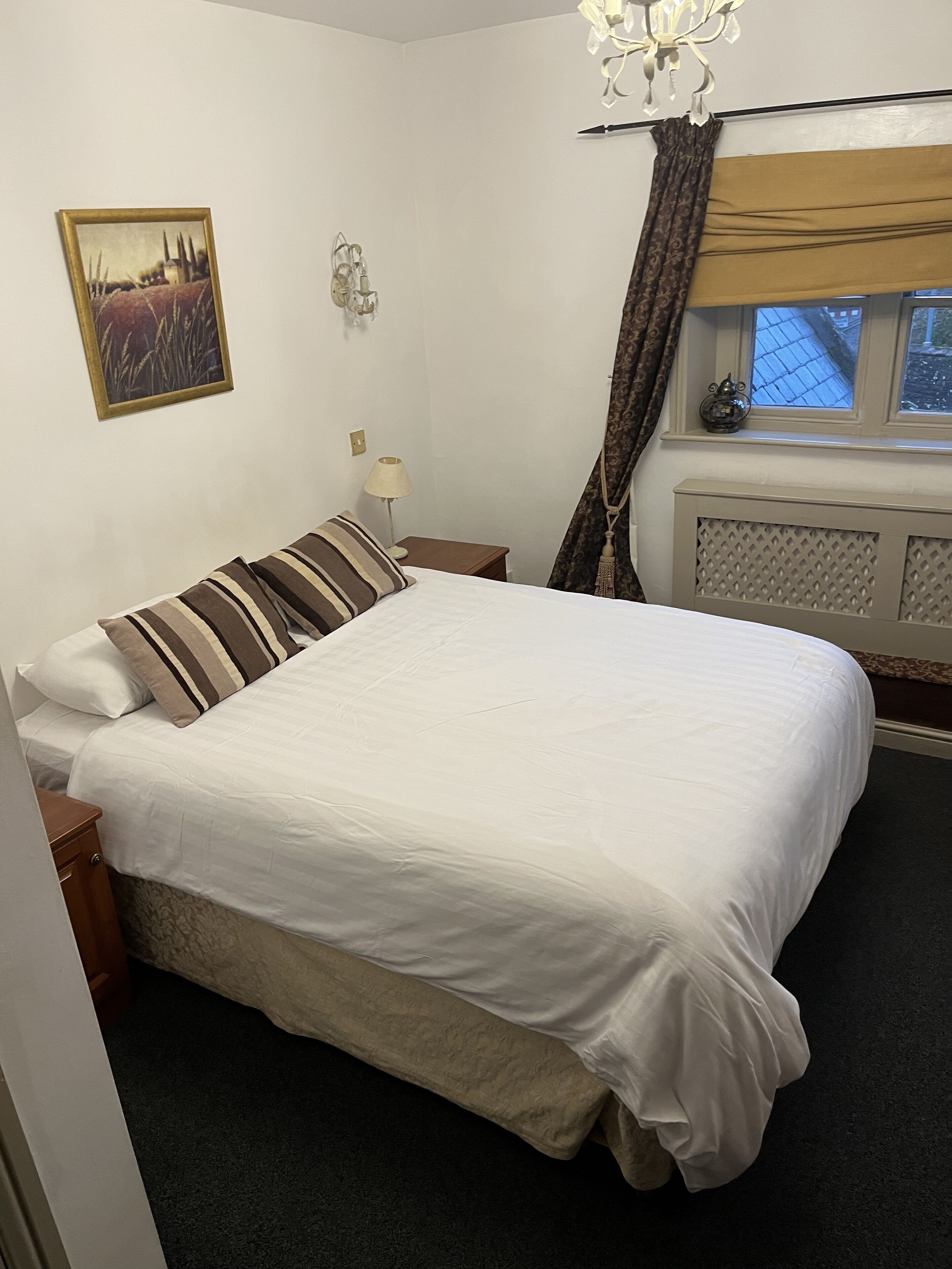 A neatly made bed with a white comforter and striped pillows, a bedside lamp, a wall-mounted light fixture, and a window with curtains.