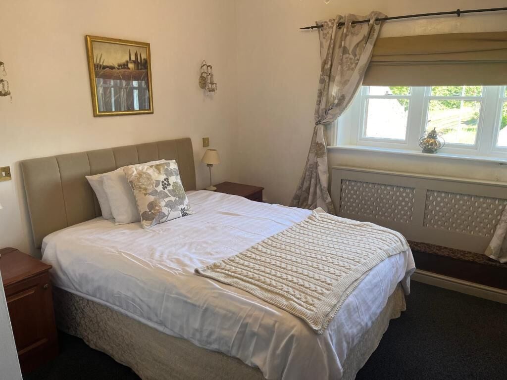 Quarto com cama, mesas de cabeceira, janela com cortinas, radiador e um quadro na parede.