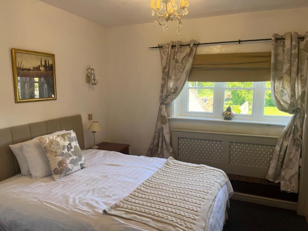 Een slaapkamer met een bed, een raam met gordijnen, een nachtkastje, een lamp en een aan de muur bevestigd schilderij.