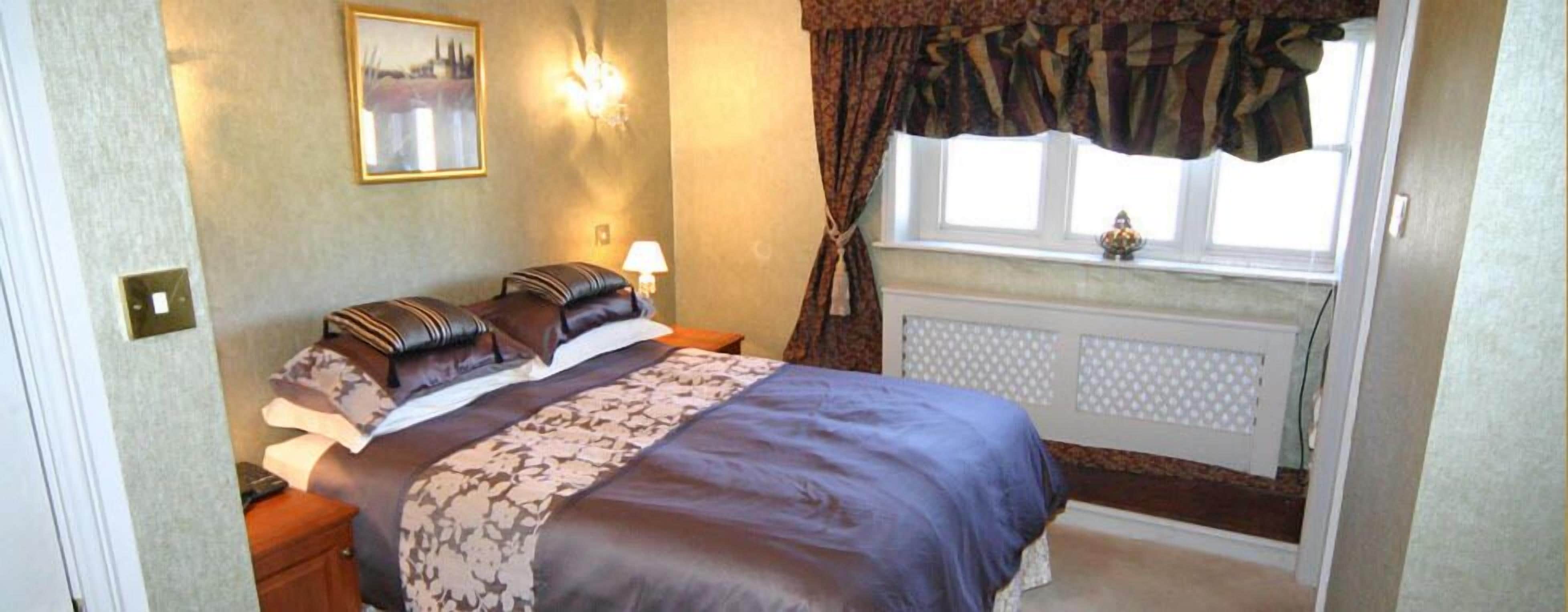 A bedroom with a bed, a nightstand, a wall-mounted lamp, a window with curtains, and a radiator.