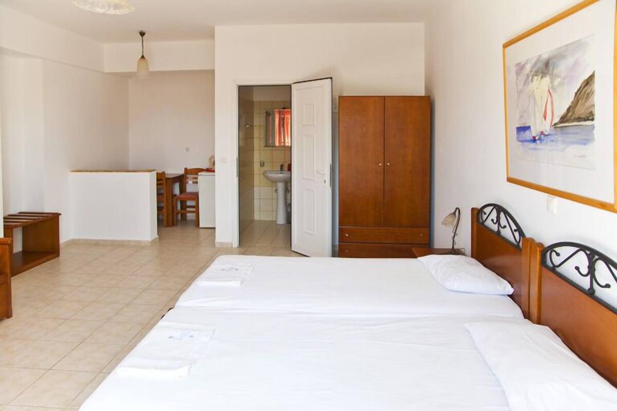 A hotel room with a large bed, a wooden wardrobe, a desk with chairs, and a bathroom visible through an open door.