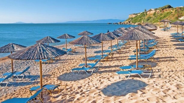 Une plage bordée de rangées de chaises longues bleues et d’ombrelles en chaume, un ciel dégagé et une mer calme.