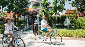 Dos personas montando en bicicleta por un camino pavimentado, con un edificio y palmeras al fondo.