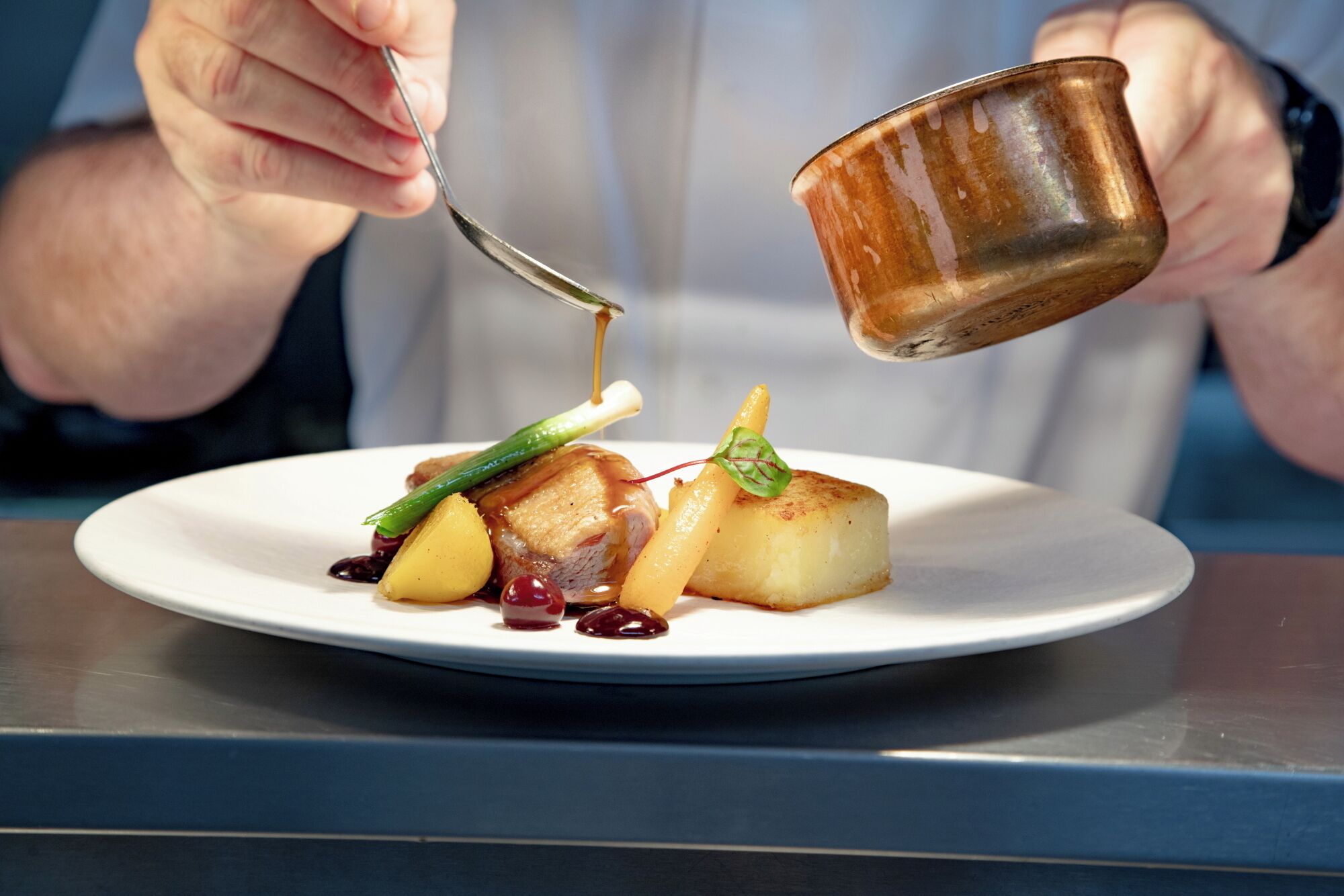 A person pouring a sauce from a small metal cup onto a plate with food.