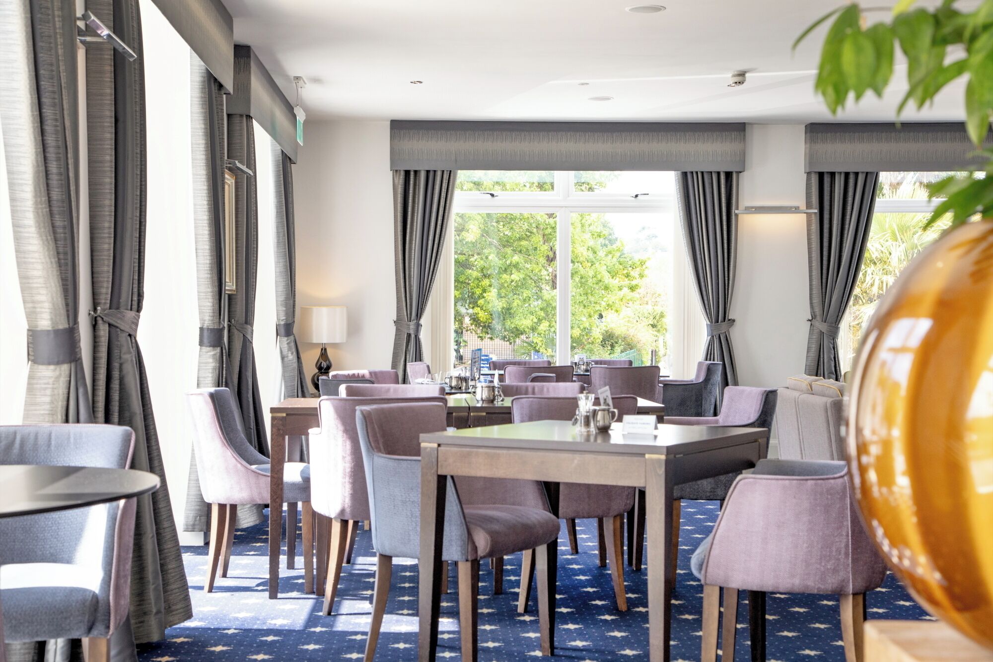Comedor de hotel con mesas, sillas, ventanas amplias y cortinas.