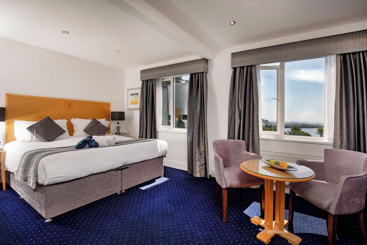 A hotel room with a bed, two chairs, a small table with fruit, and a window with a view.