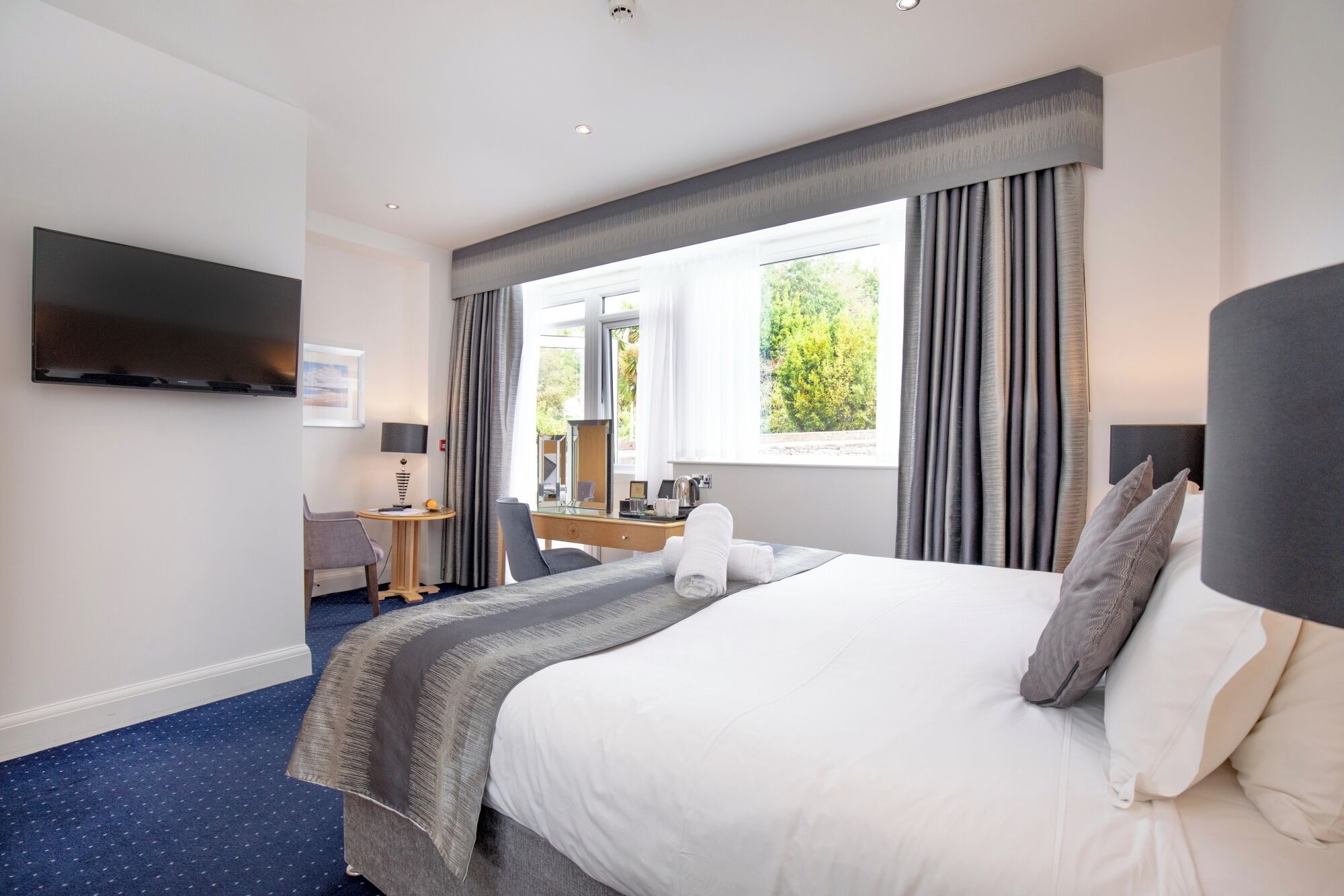 A hotel room with a bed, a desk, a chair, a flat-screen TV, and a window with curtains.