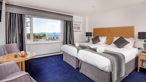 A hotel room with a large bed, two bedside tables, a small round table with a remote control and fruit, and a view of the sea. - Belgrave Sands Hotel & Spa (Torquay)