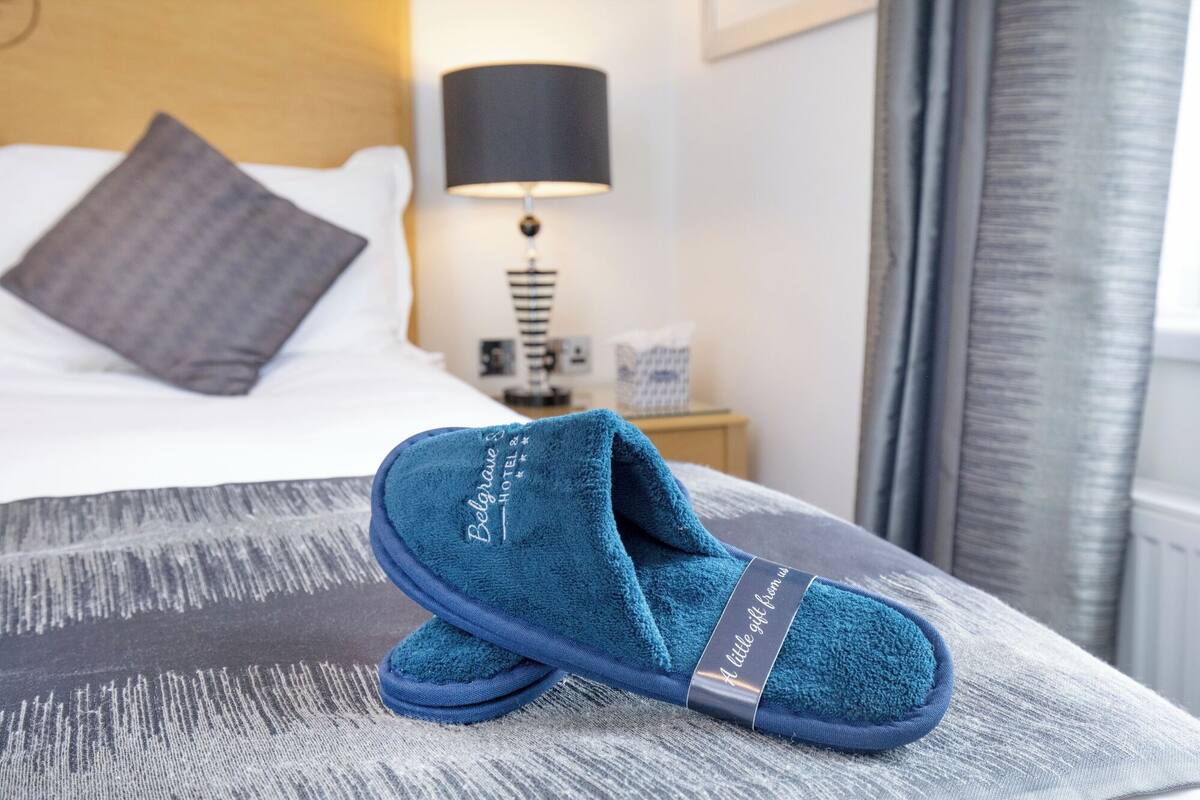 A bed with a patterned bedspread, a pillow, a nightstand with a lamp, and a pair of slippers with a label.