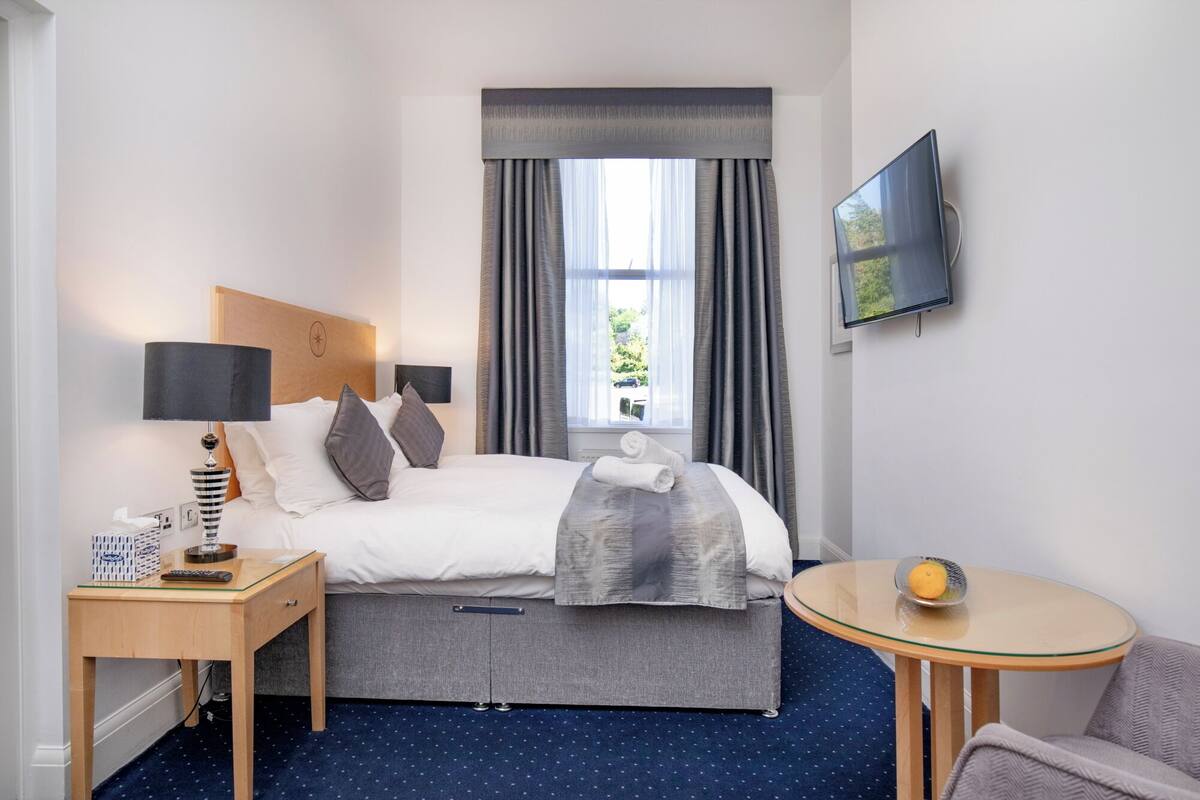 A hotel room with a bed, bedside table, lamp, TV, and a small round table with an orange.