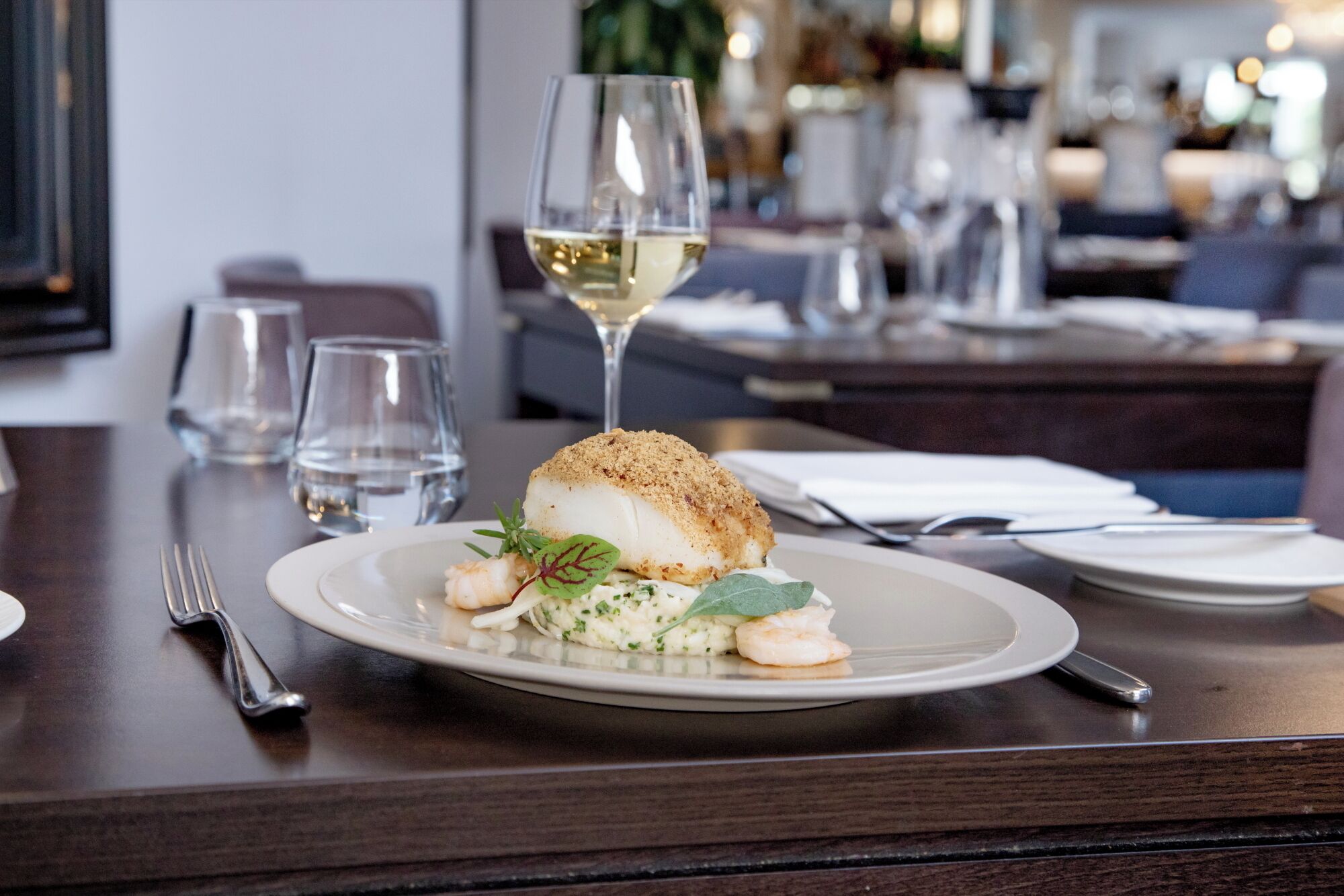 A dining table with a plate of food, a glass of white wine, and cutlery.
