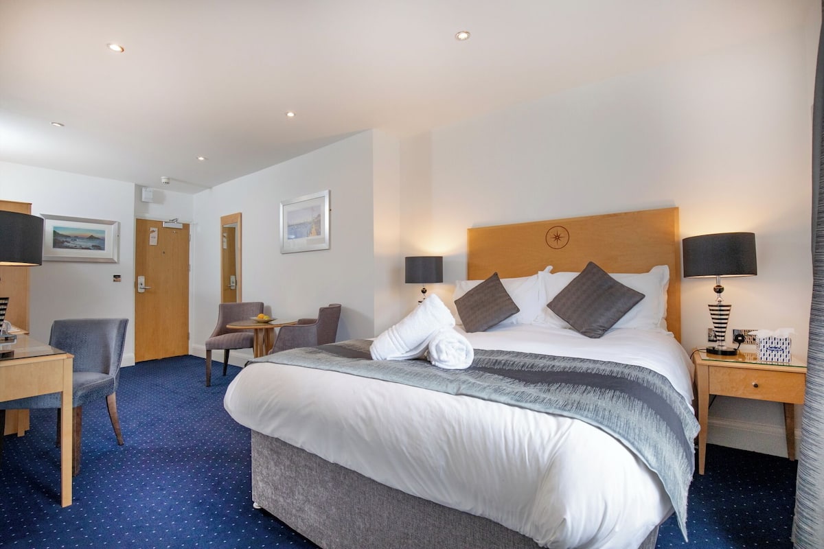 A hotel room with a bed, two bedside tables, a desk with a chair, a small table with chairs, and a door.