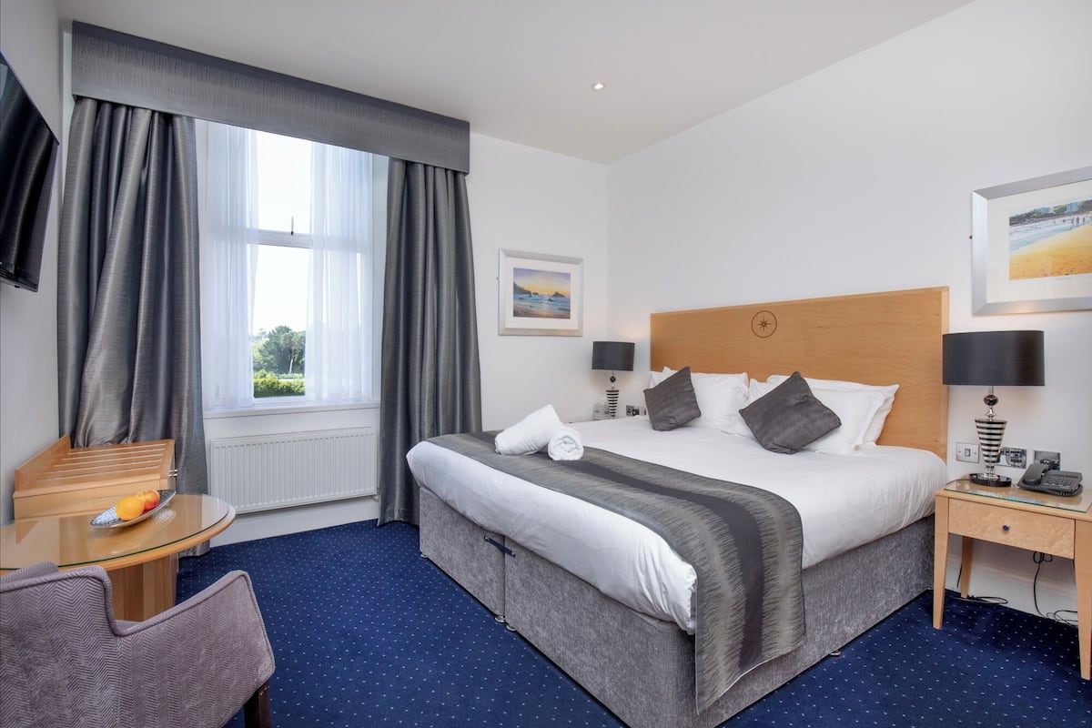 A hotel room with a bed, a desk, a chair, a window with curtains, and artwork on the walls.