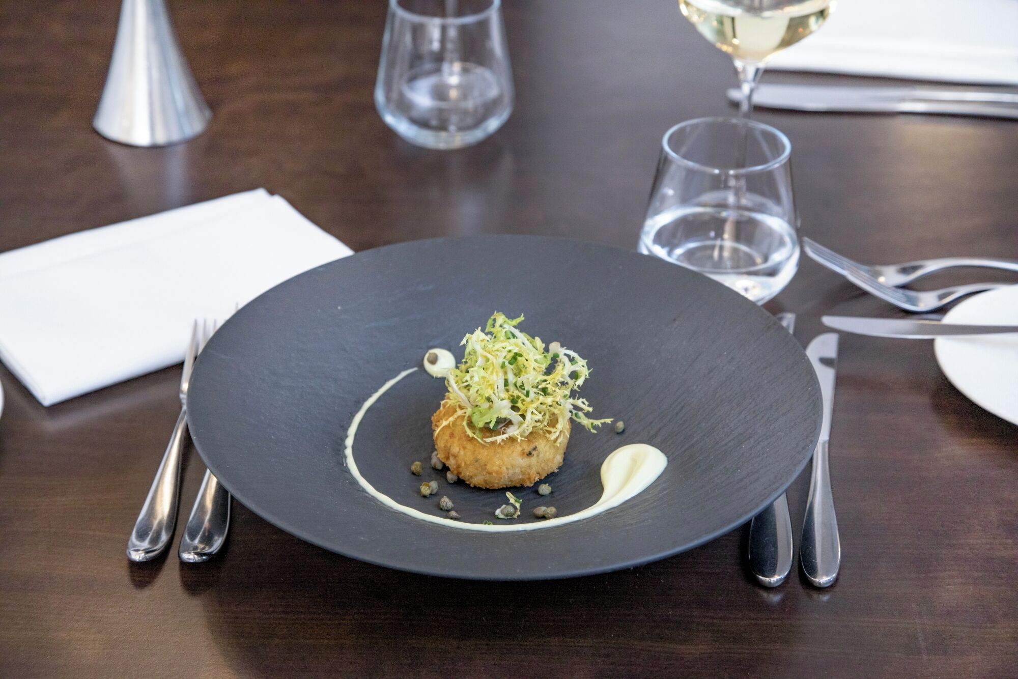 A black plate with a food item, silverware, a glass of white wine, and a napkin on a wooden table.