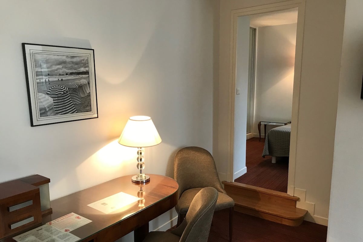 A hotel room with a wooden desk, a lamp, a framed picture on the wall, a chair, and a mirror reflecting the room.