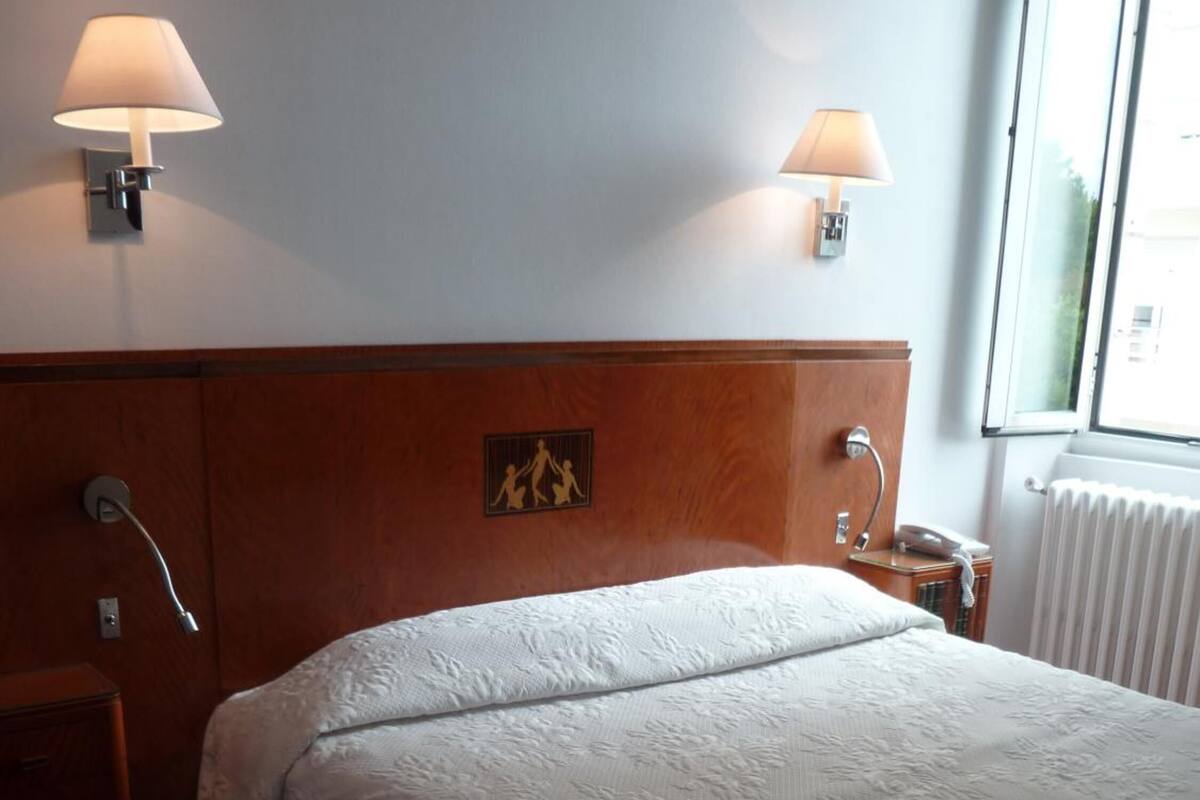A hotel room with a large bed, two wall-mounted lamps, a wooden headboard, a small bedside table with a lamp, and a radiator.