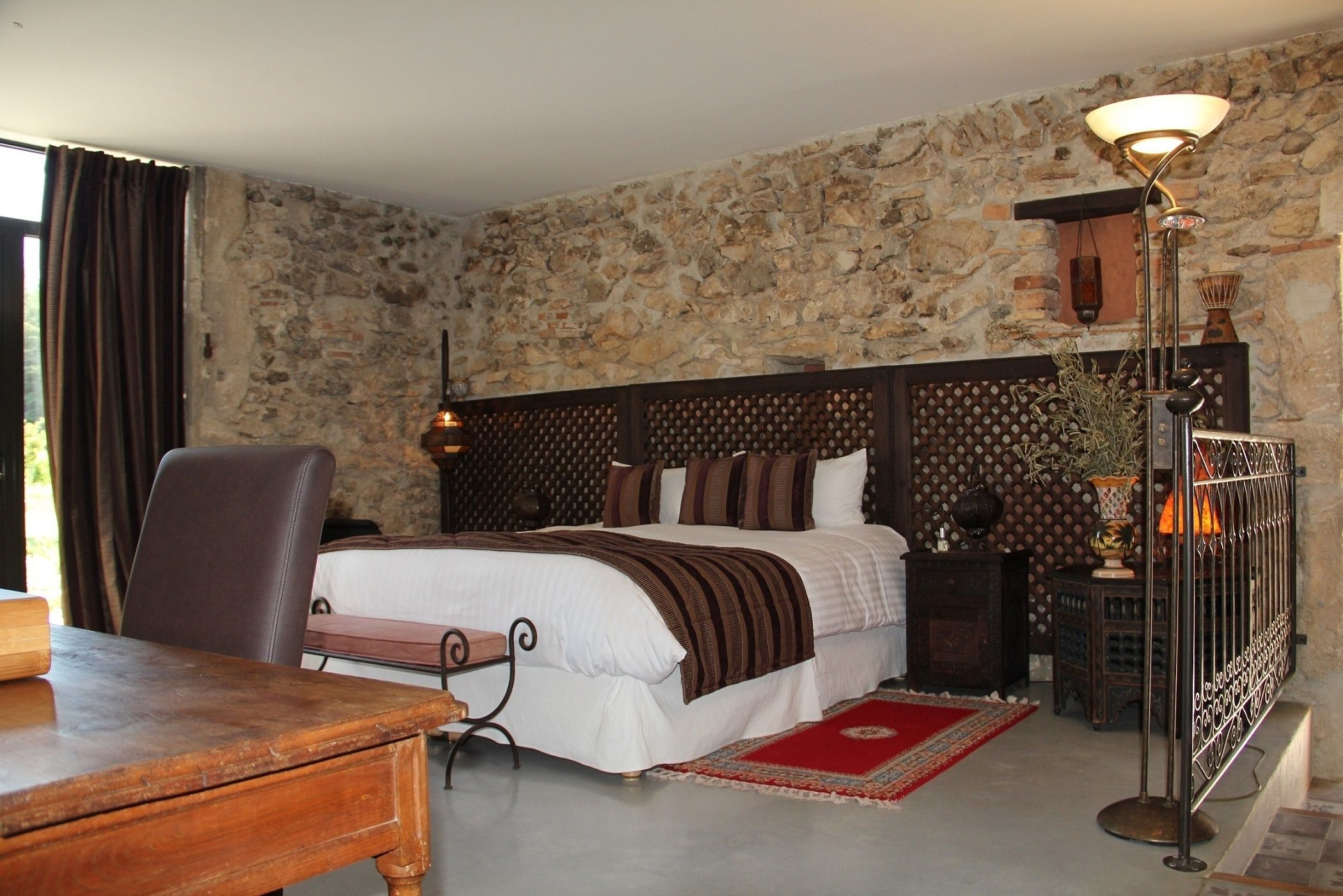 A bedroom with a stone wall, a bed with a wooden headboard, a desk, a chair, a lamp, and a red rug.