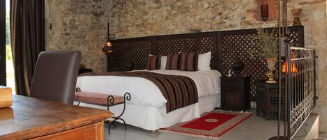 A bedroom with a stone wall, a bed with a wooden headboard, a desk, a chair, a lamp, and a red rug.