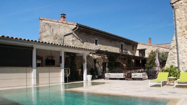Una piscina con agua cristalina, un edificio de piedra con techo de tejas y mobiliario exterior con sillas y una mesa.