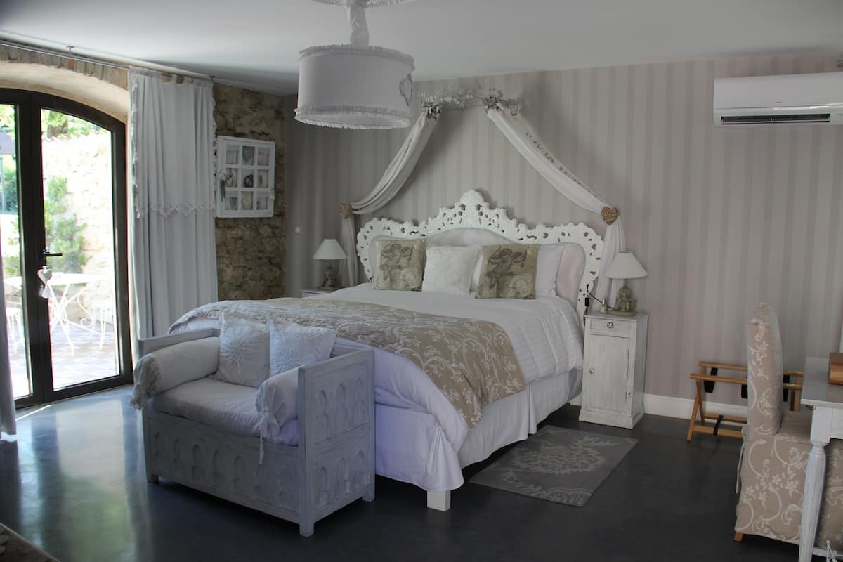 Un dormitorio con una cama grande, una mesita de noche blanca, una silla de madera y una ventana con cortinas.