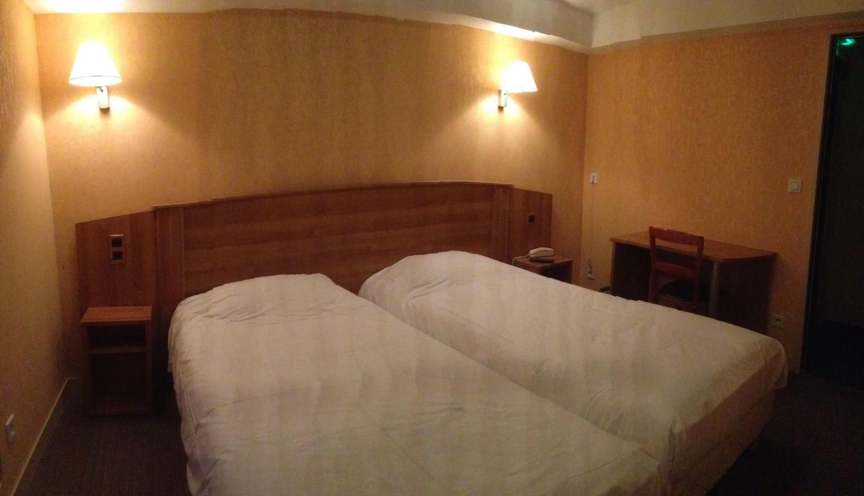 Una habitación de hotel con una cama doble, dos mesitas de noche, un cabecero de madera y dos lámparas fijadas a la pared.