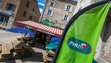 A restaurant with outdoor seating, a sign for 'Hotel de l'Abbaye', and a 'PMUS' logo on a green banner.