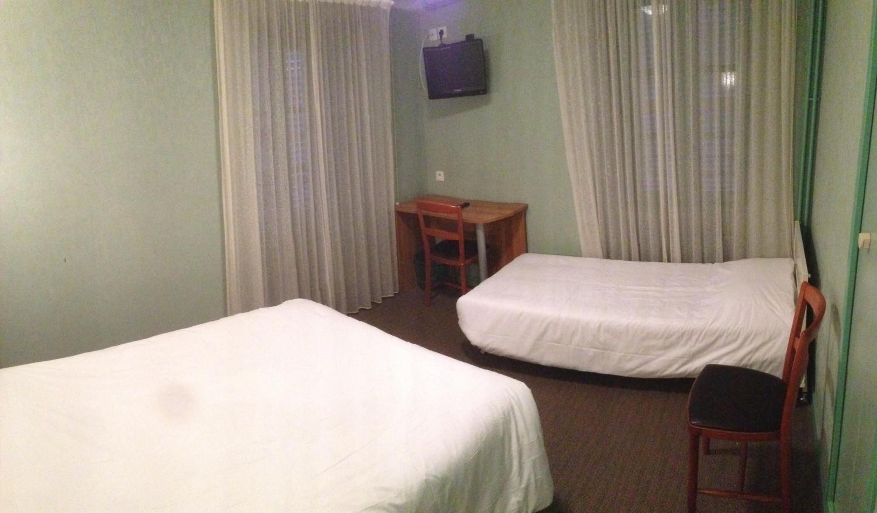 A small hotel room with two single beds, a desk, a chair, and a television on the wall.