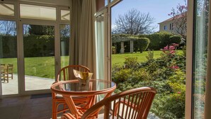 Um alpendre envidraçado com uma mesa redonda e cadeiras, vista para um jardim bem cuidado, e um edifício ao fundo.