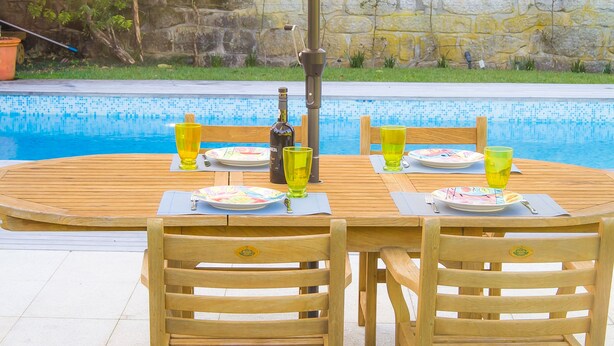 Uma mesa de madeira com cadeiras junto a uma piscina, com pratos, copos e uma garrafa sobre a mesa.