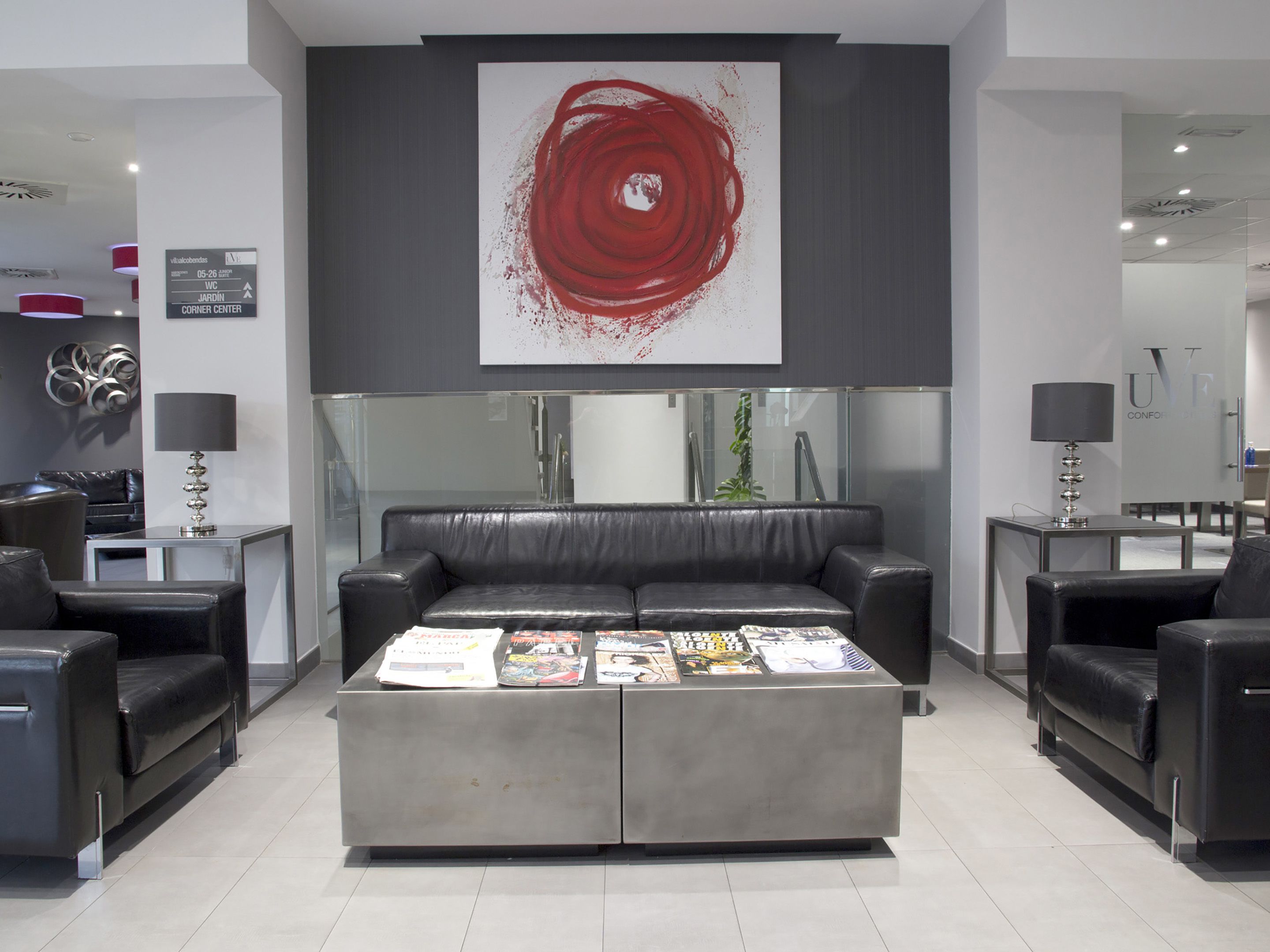 Un hall d’entrée moderne avec un canapé central de couleur grise, des fauteuils en cuir noir et une table basse sur laquelle sont posés des magazines. Une grande peinture abstraite rouge est accrochée au-dessus du canapé.