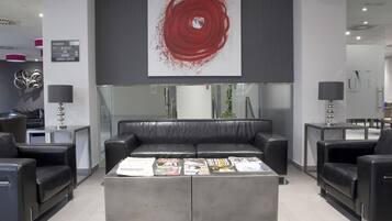 Un hall d’entrée moderne avec un canapé central de couleur grise, des fauteuils en cuir noir et une table basse sur laquelle sont posés des magazines. Une grande peinture abstraite rouge est accrochée au-dessus du canapé.