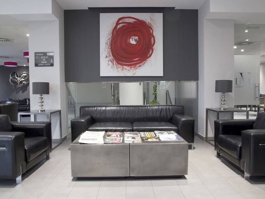 Un hall d’entrée moderne avec un canapé central de couleur grise, des fauteuils en cuir noir et une table basse sur laquelle sont posés des magazines. Une grande peinture abstraite rouge est accrochée au-dessus du canapé.