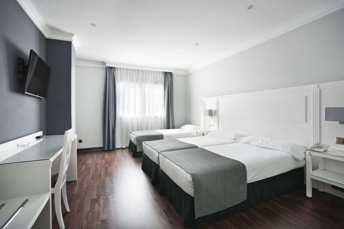 Una camera d'albergo con una scrivania, due letti, una televisione e una finestra con tende.