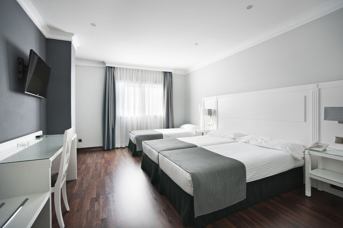 Habitación de hotel con escritorio, dos camas, televisión y ventana con cortinas.