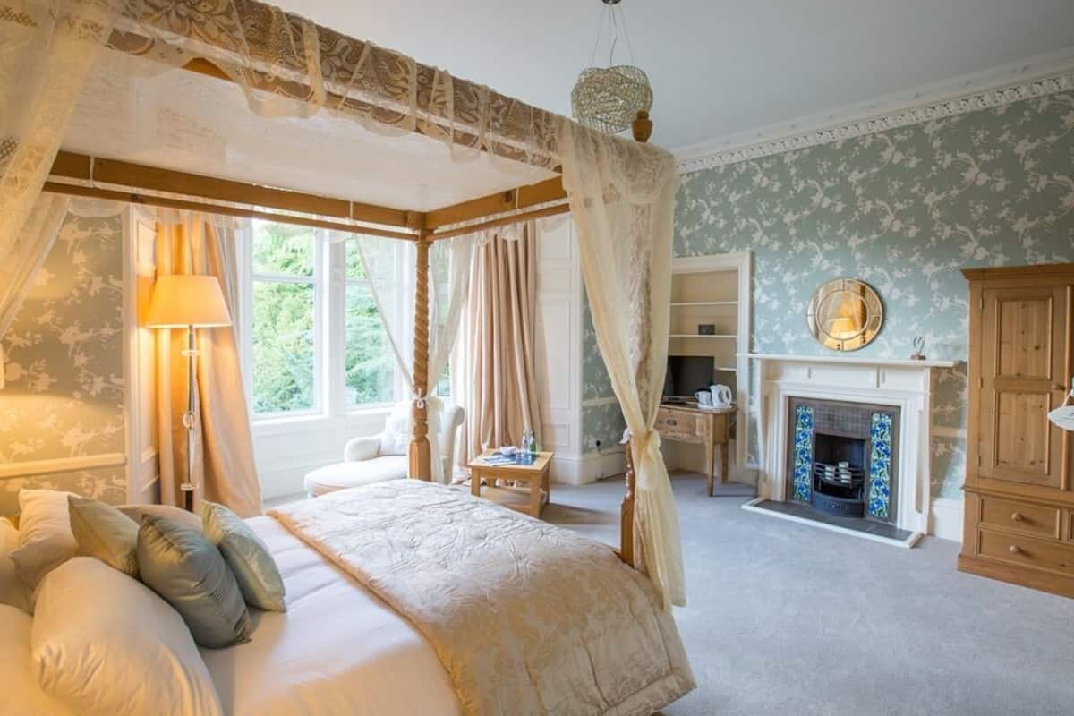 A bedroom with a four-poster bed, a fireplace, and a view of greenery outside.