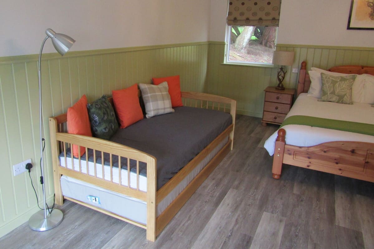 A room with two wooden beds, one with a headboard and one without. There is a window with a valance, a bedside table with a lamp, and a floor lamp.