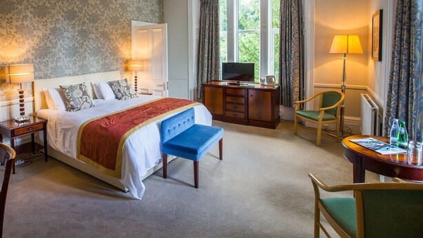 A hotel room with a large bed, a desk with a TV, a chair, and a table with a lamp and bottles. - Balcary House Hotel (Hawick)