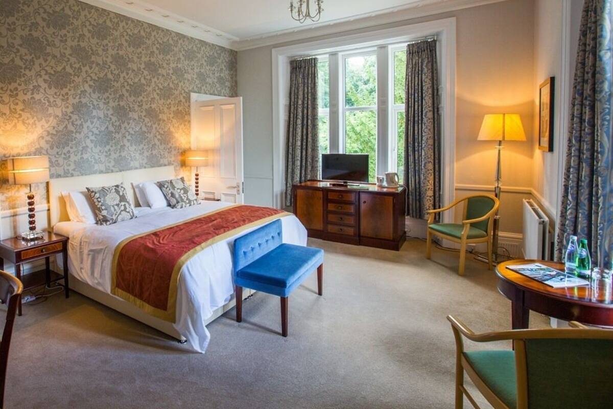 A hotel room with a large bed, a desk with a TV, a chair, and a table with a lamp and bottles.