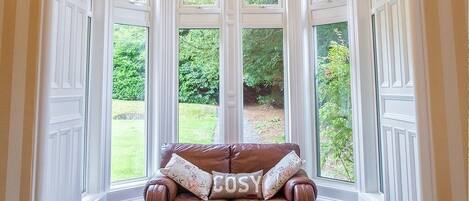 A corner room with a brown leather sofa and a window with a view of greenery.