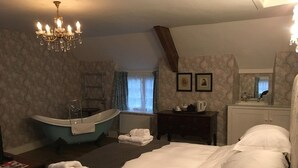 A hotel room with a large bed, a bathtub, a vanity with a mirror, and a chandelier. - Clare Cottage (Sherborne)