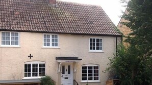 A two-story building with a traditional design, featuring a tiled roof, a front door with a small porch, and a cross symbol above the door. - Clare Cottage (Sherborne)