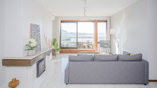 Un soggiorno moderno con un divano grigio, un caminetto, una TV e vista sulla città attraverso ampie finestre.