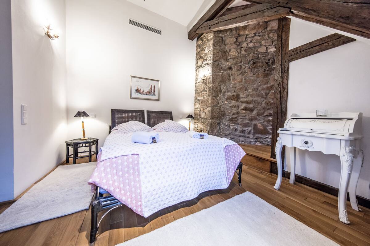 Une chambre à coucher comprenant un lit, une table de chevet, une commode, un mur en pierre et une poutre en bois au plafond.