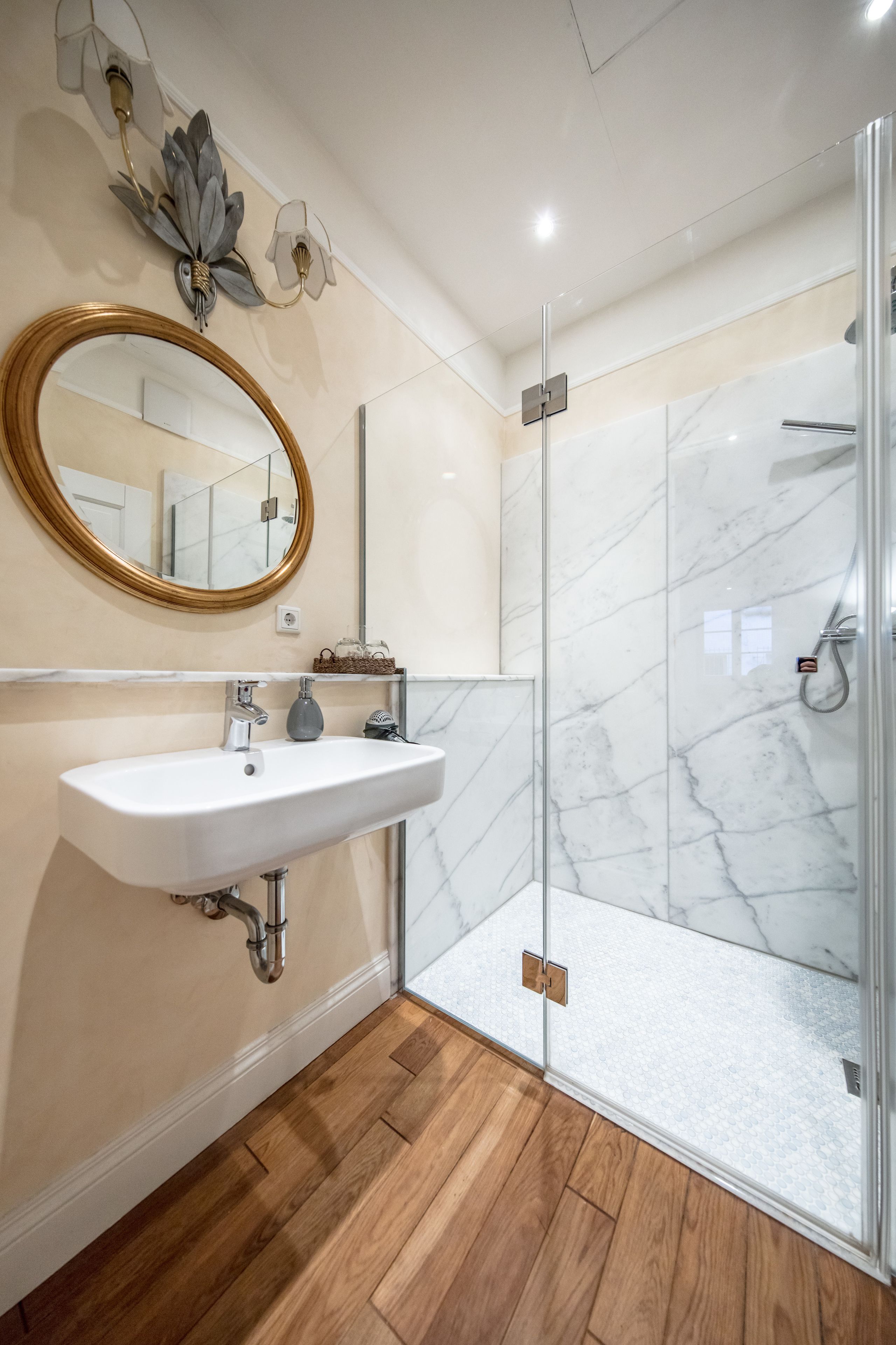 Une salle de bain avec une douche en marbre, un plancher en bois et un lavabo blanc agrémenté d’un miroir doré.