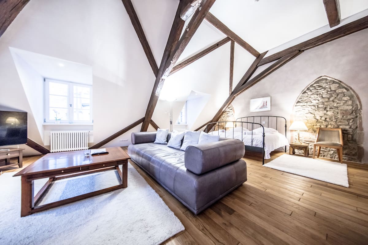 Une chambre à coucher de style loft avec des poutres en bois apparentes, un canapé gris, un lit à baldaquin blanc, un mur en pierre et une vue sur l’extérieur.