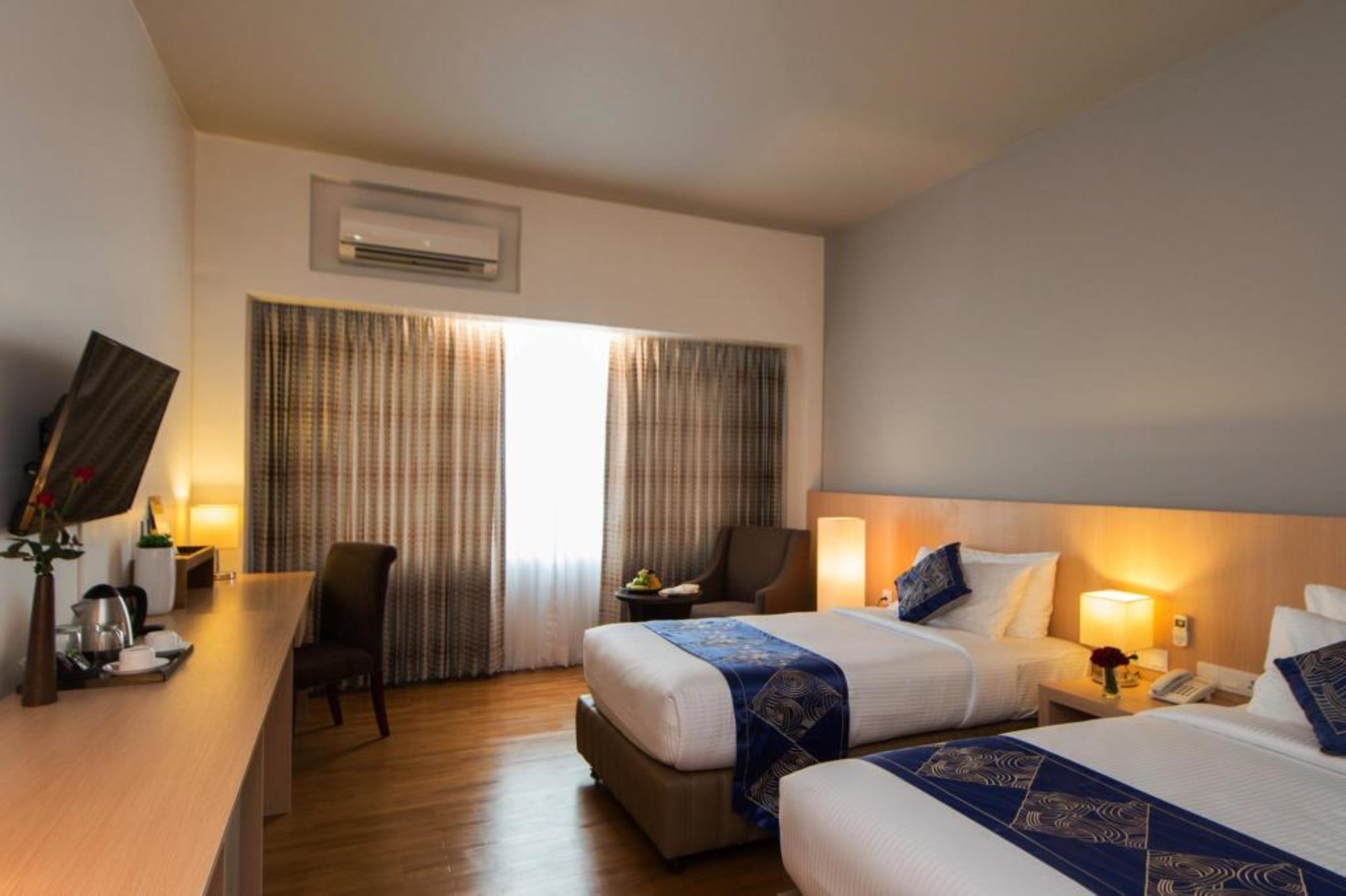 Quarto de hotel com duas camas, uma escrivaninha, uma cadeira, uma televisão e uma janela com cortinas.